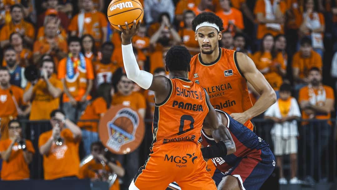 TJ Shorts, con la camiseta naranja número 0 del Valencia, levanta la pelota para lanzar ante un defensor del Baskonia, con la tribuna naranja detrás