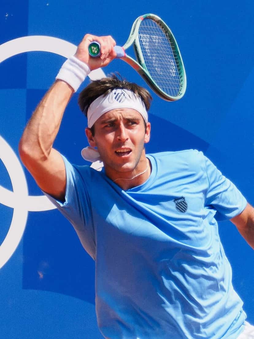 Foto de archivo: Tomás Martín Etcheverry, con remera celeste y vincha blanca, le pega un drive con los anillos olímpicos en la pared del fondo