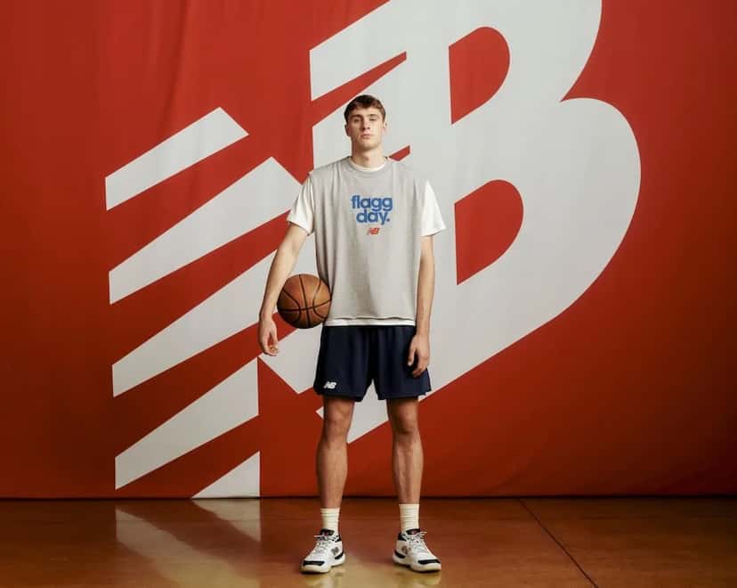 Cooper Flagg parado sobre un piso de parquet frente a una pared roja con el logo gigante de New Balance en blanco, con una pelota de básquet bajo el brazo, chaleco gris con la frase flagg day en azul, short azul marino y zapatillas blancas; foto de archivo de la campaña Flagg Day