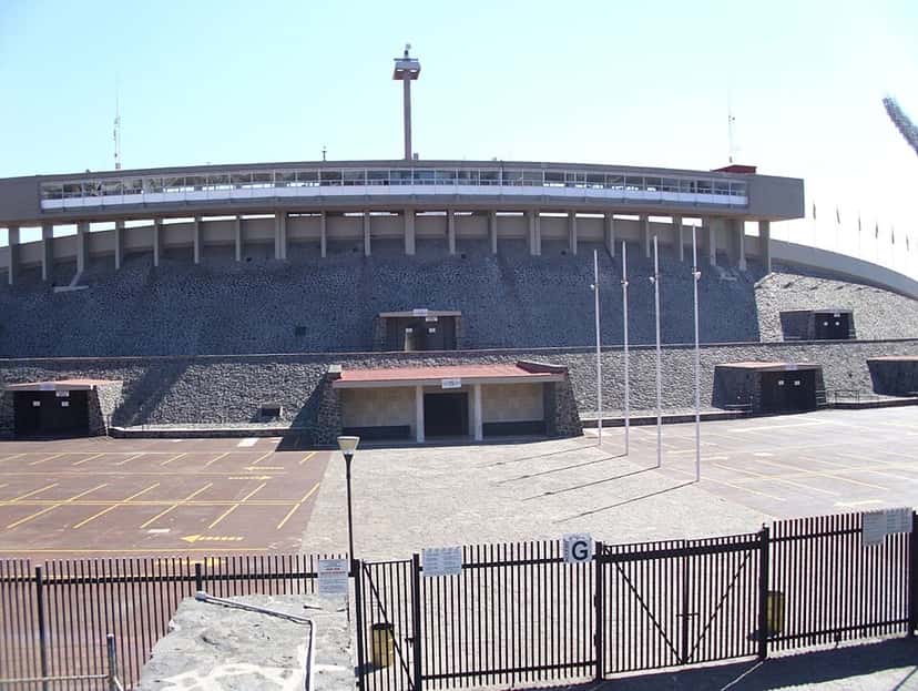 La fachada del estadio Olímpico Universitario, con su tribuna de piedra y los mástiles en la explanada de acceso; foto de archivo de 2011 (foto: Ebidej, Wikimedia Commons, licencia CC BY-SA 3.0)