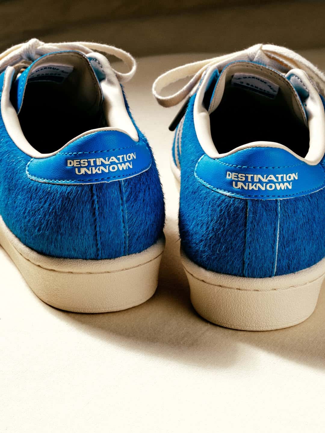 Talones de la adidas Superstar de Liberaiders, con el pelo largo azul, la pieza del talón con la frase Destination Unknown y la suela color crema