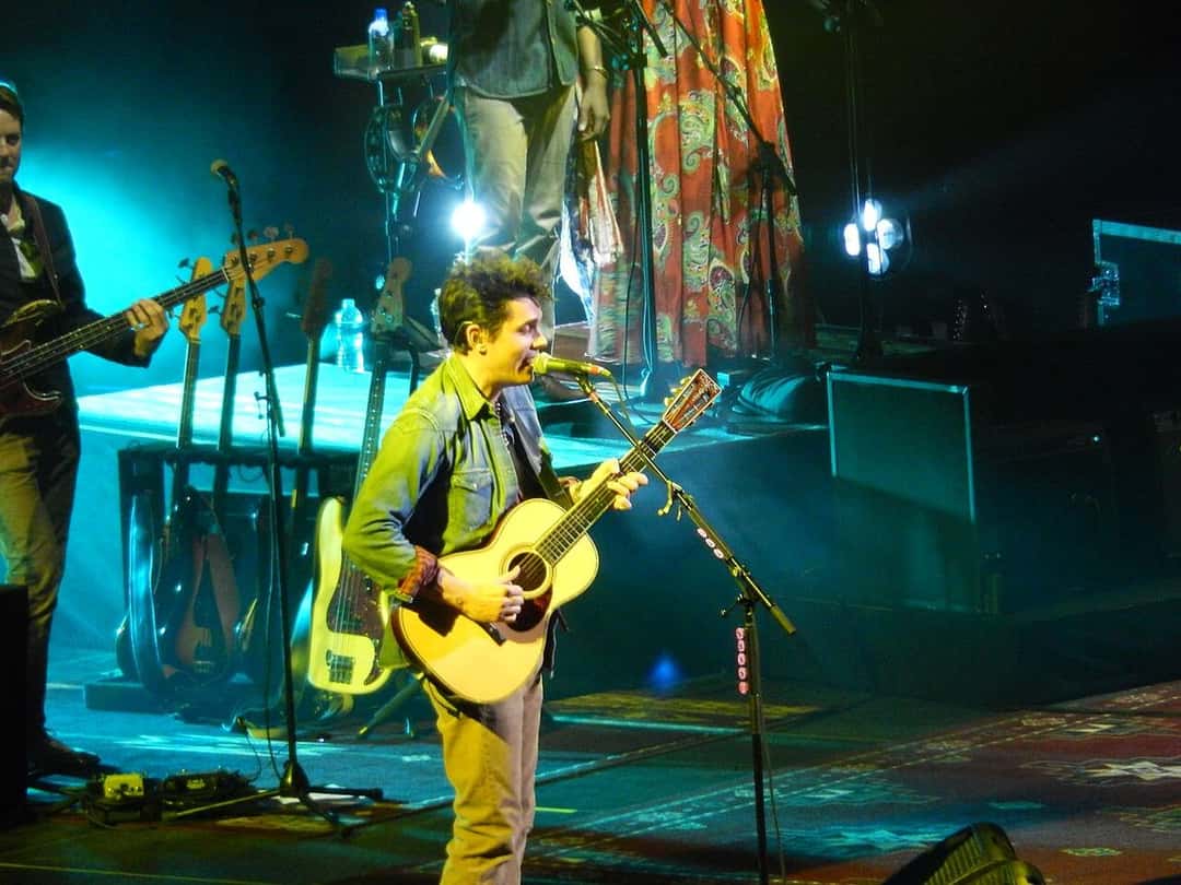Foto de archivo de John Mayer en vivo en el Barclays Center de Nueva York, en diciembre de 2013: canta frente al micrófono con una guitarra acústica y camisa de jean, bajo luces celestes. Foto: slgckgc, Wikimedia Commons, CC BY 2.0
