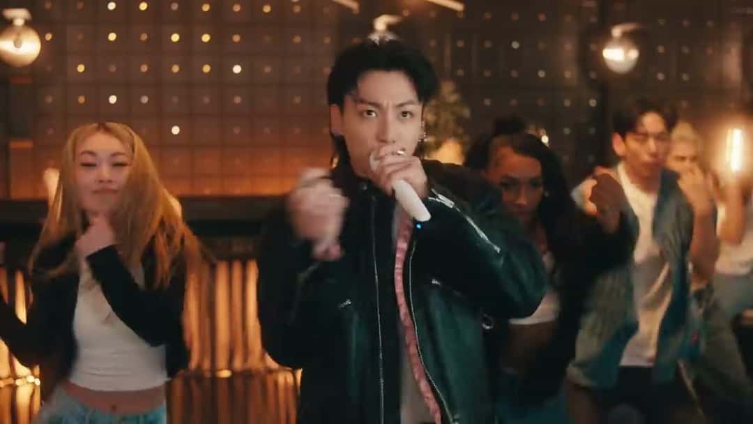 Fotograma del video oficial de la performance de «3D»: Jungkook canta con un micrófono y campera de cuero negra, rodeado de bailarines, en un salón con luces cálidas