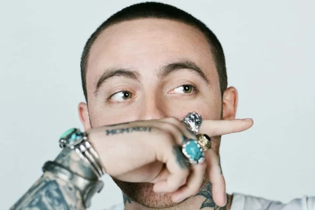 Retrato de Mac Miller sobre fondo blanco, mirando hacia un costado y tapándose parte de la cara con la mano tatuada y llena de anillos de turquesa y plata