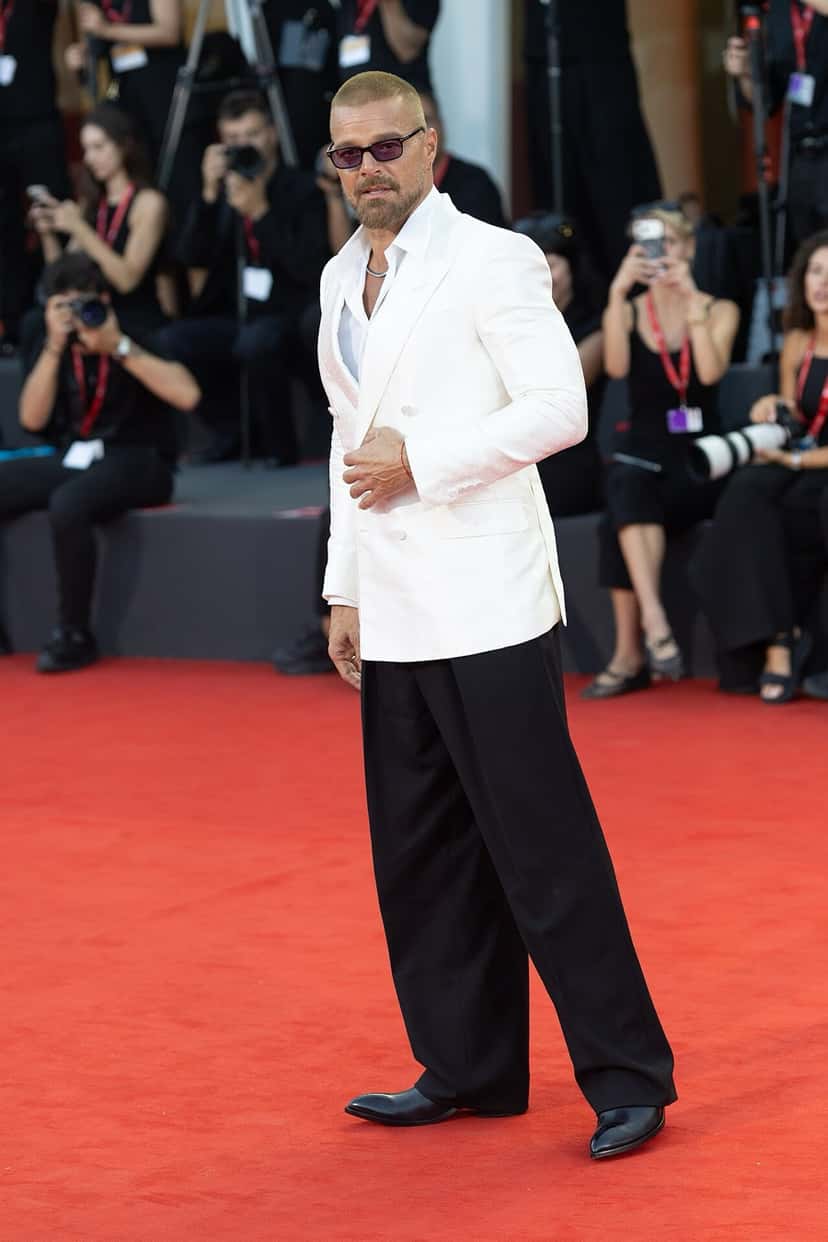 Ricky Martin en una alfombra roja con saco cruzado blanco, camisa abierta, pantalón negro ancho y anteojos de sol de cristales violetas, con fotógrafos detrás. Foto de archivo en el Festival de Venecia, septiembre de 2026. Foto: LucaFazPhoto, Wikimedia Commons, CC BY-SA 4.0