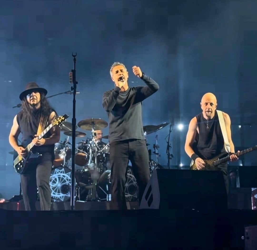 System of a Down en vivo en México en 2026 (foto de archivo): el cantante con el puño en alto en el centro del escenario, entre el guitarrista y el bajista, bajo luces azules. Foto: Californipedia, CC0, vía Wikimedia Commons