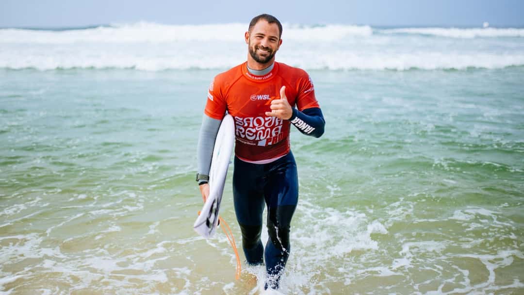 Alejo Muniz sonríe y hace el saludo surfer con la mano mientras sale del agua con su tabla bajo el brazo y la lycra naranja del Saquarema Pro (foto de archivo)