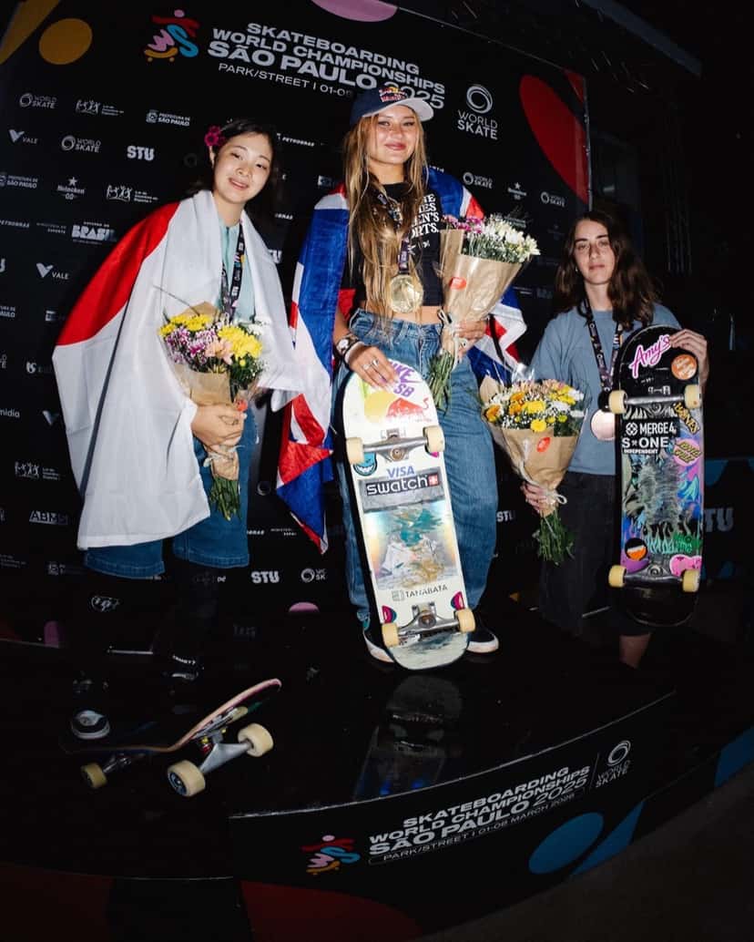 Sky Brown, envuelta en la bandera británica, sonríe con la medalla de oro, flores y su tabla en el podio del Mundial de San Pablo, con Mizuho Hasegawa a su derecha