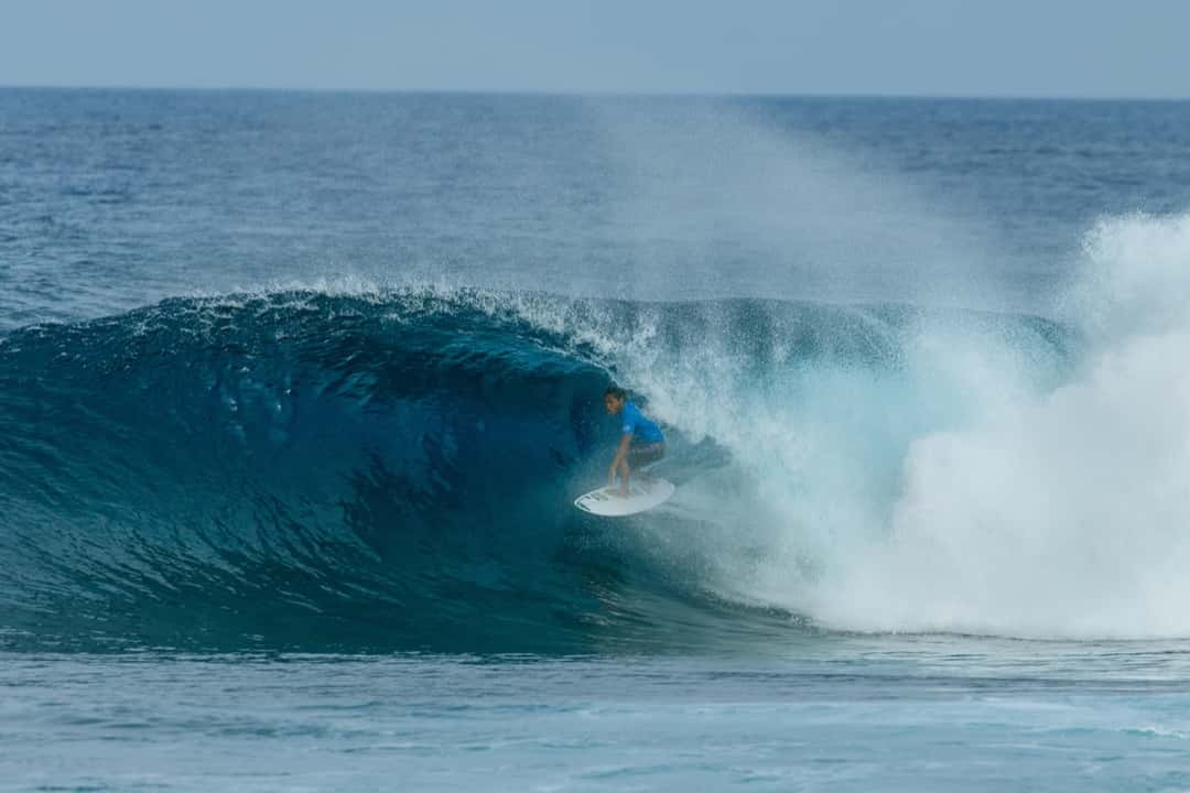 Un surfista con lycra azul agachado dentro del tubo de una ola, con la espuma que sale detrás