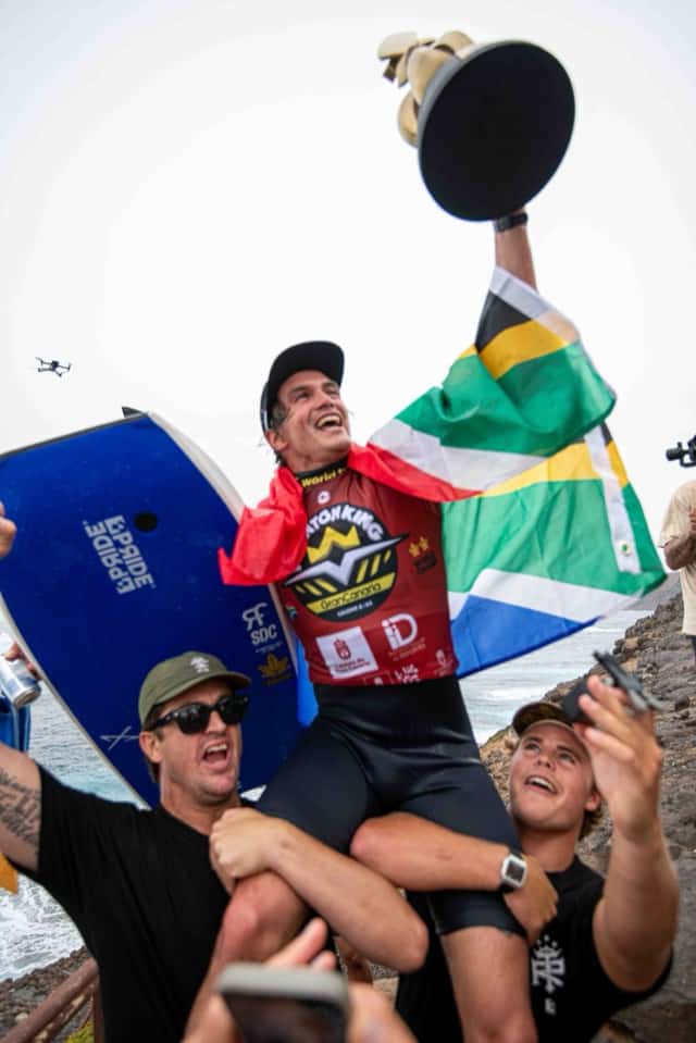 Tristan Roberts, con la camiseta roja del Frontón King y una bandera de Sudáfrica, levanta un trofeo mientras dos amigos lo llevan en andas sobre las rocas de El Frontón