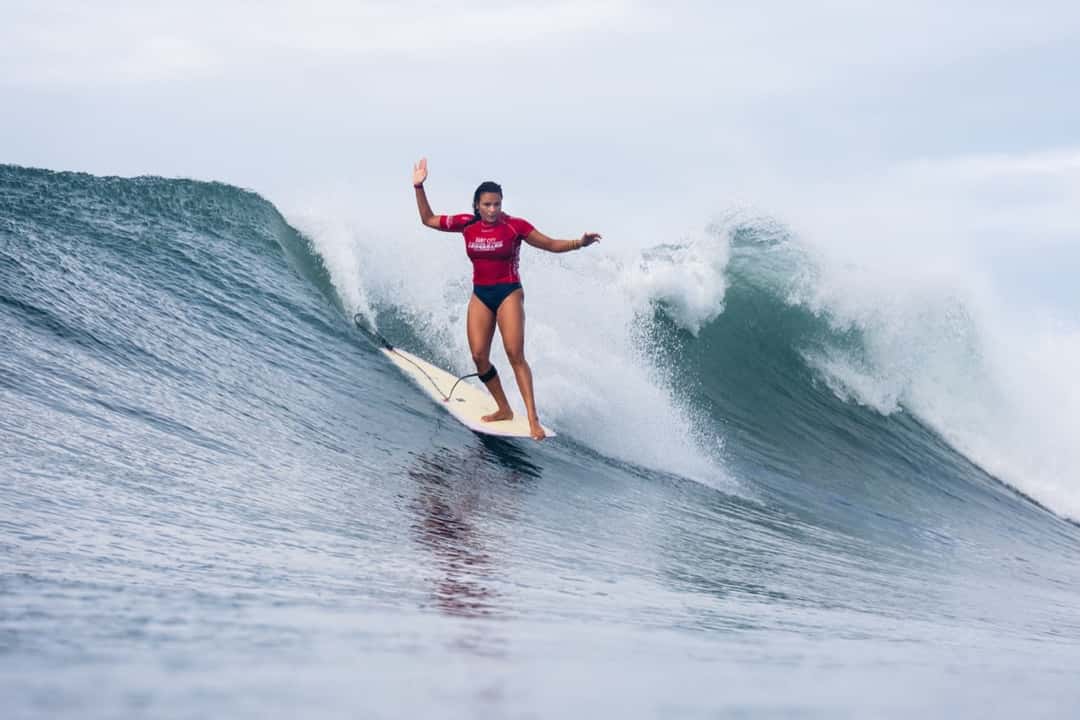Alice Lemoigne, con lycra roja, camina sobre su longboard con un brazo en alto en la pared de una ola de El Sunzal