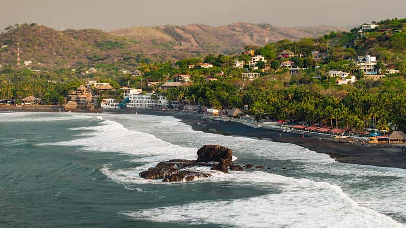 Vista de la costa de Surf City El Salvador: arena oscura, rompientes, una roca en el agua y casas entre palmeras sobre las lomas