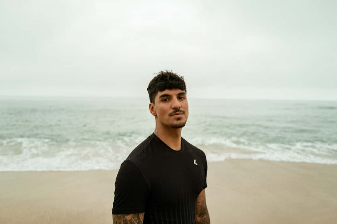 Gabriel Medina, con remera negra, mira a cámara de pie en una playa con el mar gris detrás