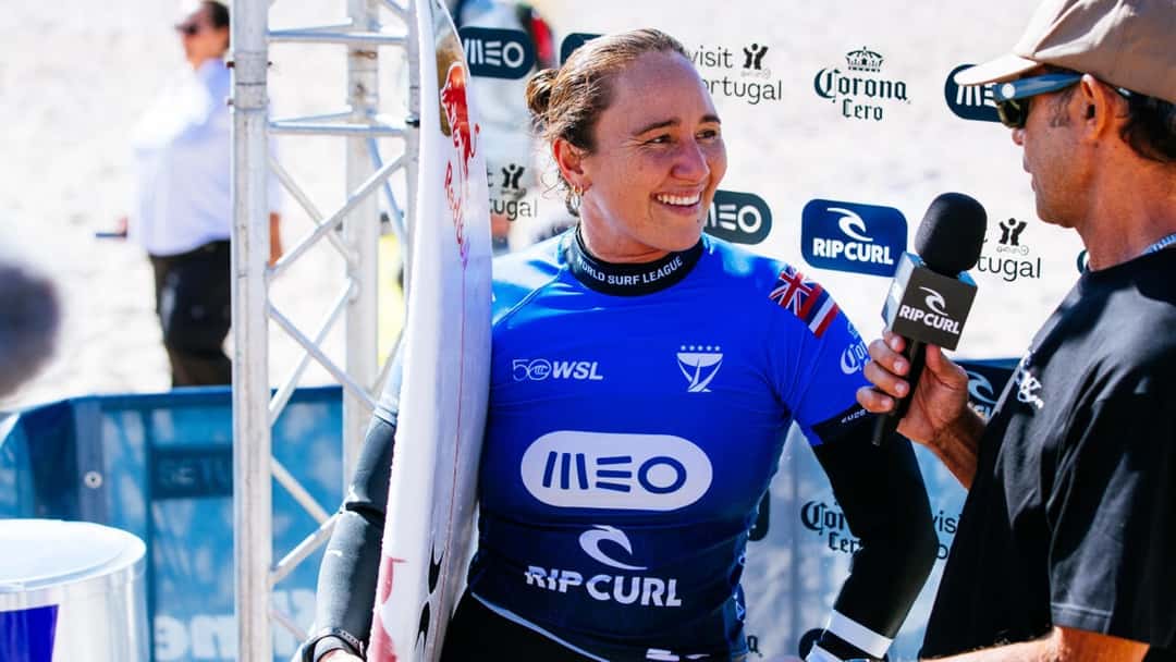 Carissa Moore, con la lycra azul del MEO Rip Curl Pro Portugal y la bandera de Hawái, sonríe con su tabla mientras la entrevistan frente al panel del evento en Supertubos
