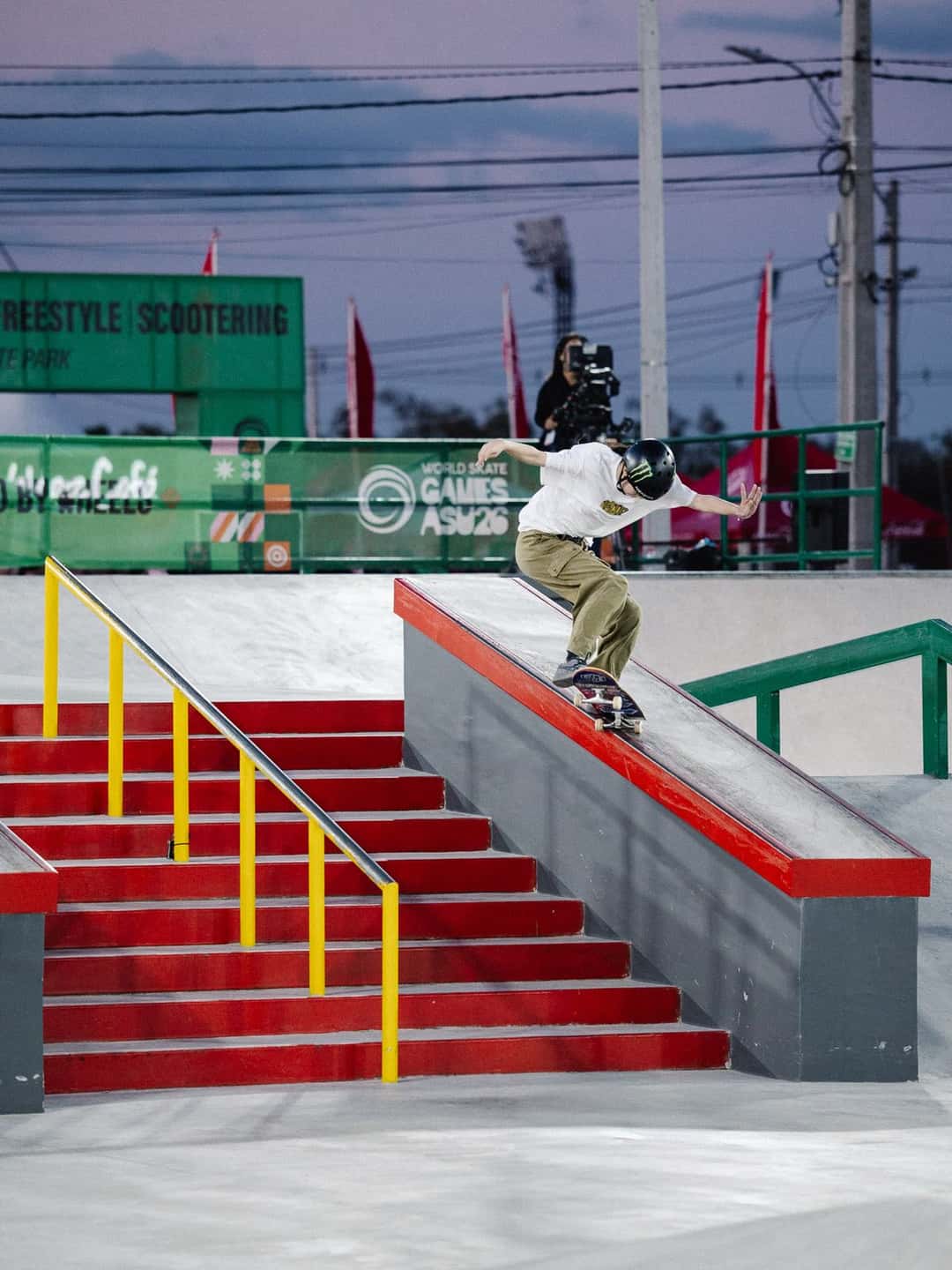 Liz Akama, con casco negro y remera blanca, se desliza sobre el borde de un cajón rojo de la pista de street de Luque en el atardecer, con un camarógrafo detrás de la valla