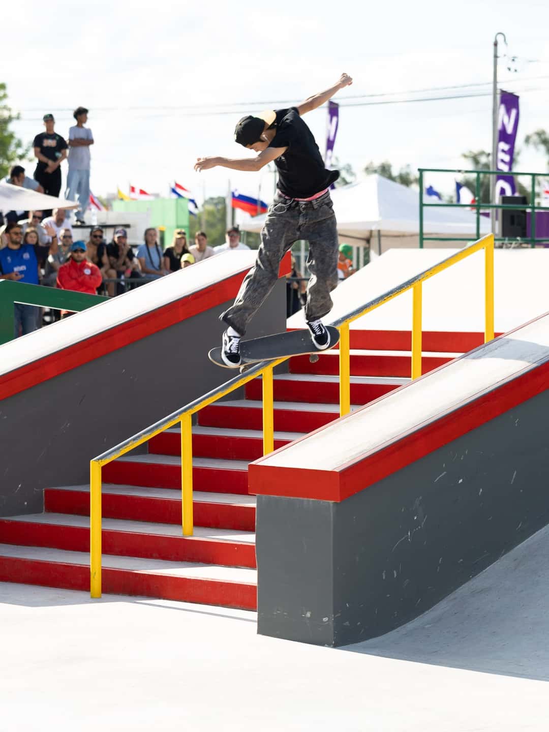 Toa Sasaki, con remera negra y gorra, desliza sobre la baranda amarilla de la escalera roja de la pista de street de Luque, con público y banderas detrás