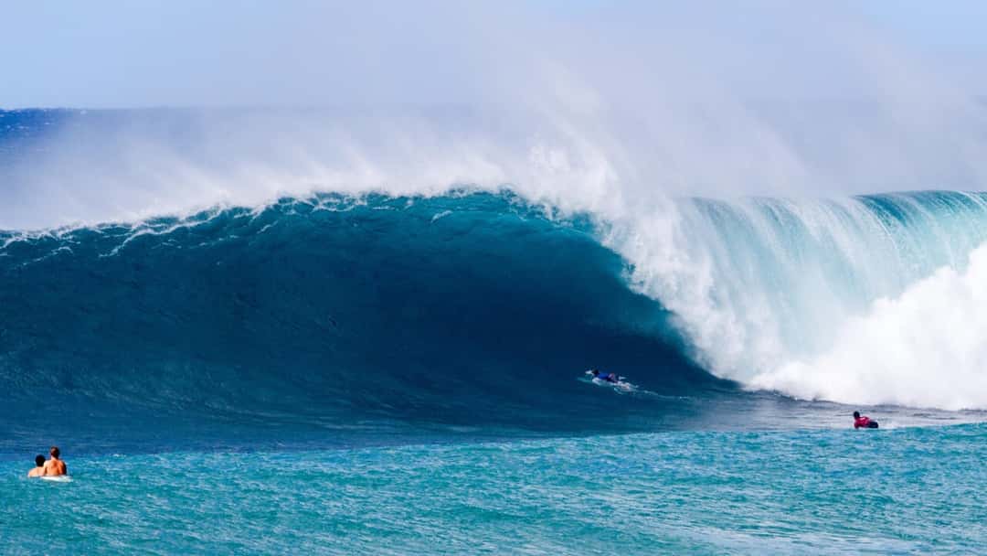 Una ola enorme rompe en tubo en el North Shore de Oahu frente a dos surfistas que reman en el canal