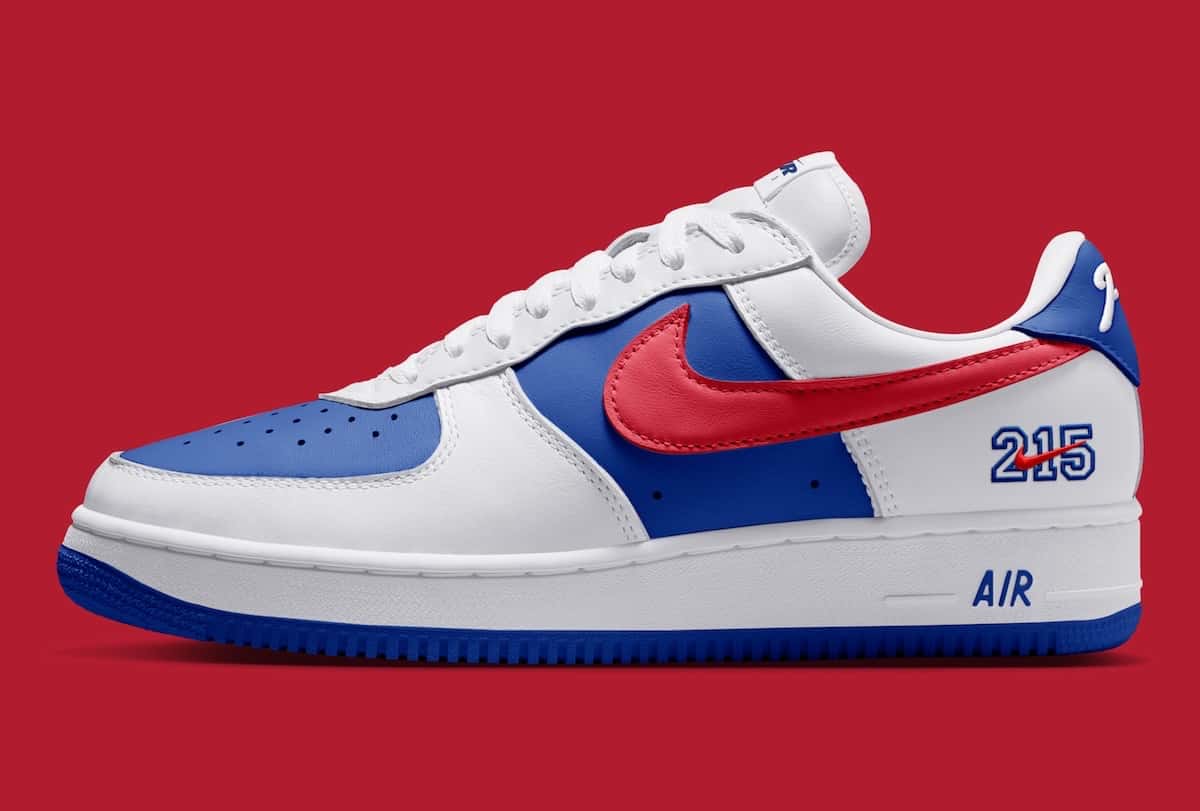 Nike Air Force 1 '01 Philly de perfil sobre fondo rojo, con cuero blanco, paneles azules, Swoosh rojo, el número 215 en el talón y la palabra AIR en azul sobre la mediasuela