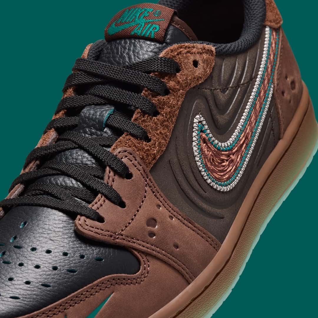 Primer plano del lateral del Air Jordan 1 Low OG Somos Historias, con los cordones negros, la gamuza marrón de pelo largo en los ojales, el cuero marrón oscuro con relieves en ondas, el swoosh tejido marrón con borde verde agua y gris, y el logo Nike Air verde en la lengüeta