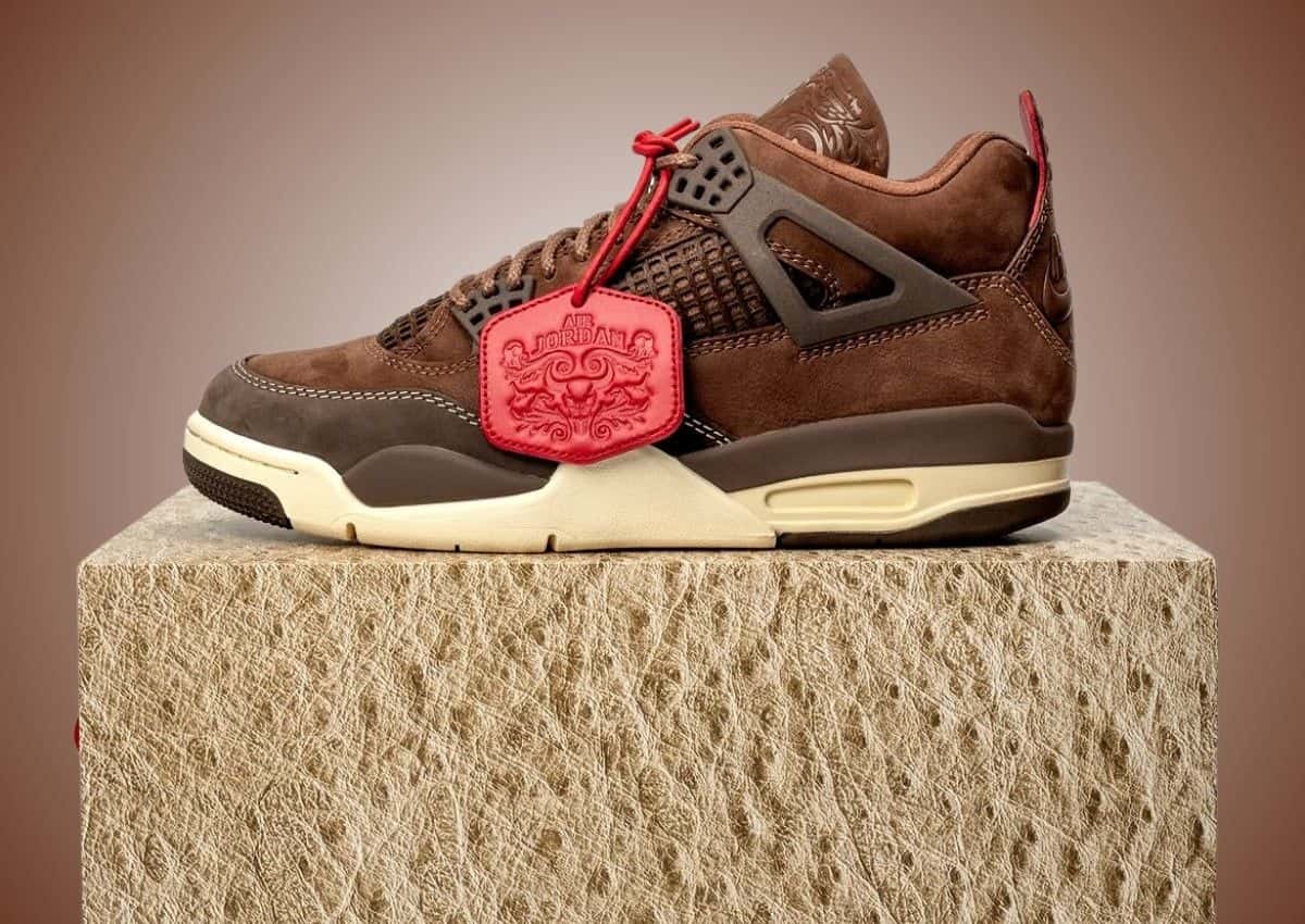 Air Jordan 4 BIN 23 en gamuza marrón con colgante de cuero rojo, apoyado sobre una caja con textura de avestruz