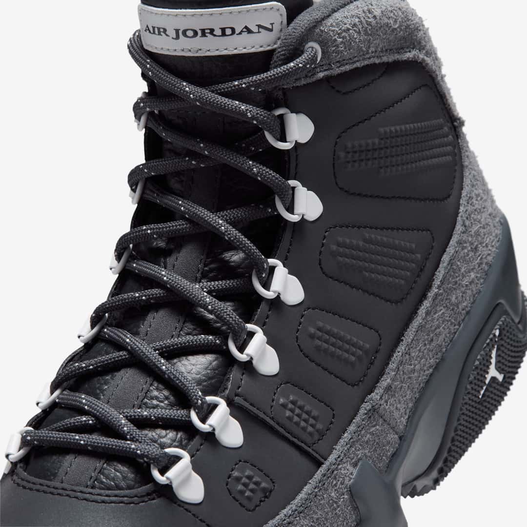 Primer plano del Air Jordan 9 Retro Boot NRG Anthracite mostrando los cordones gris oscuro con puntos claros pasando por los ojales metálicos en forma de D con terminales grises, la etiqueta Air Jordan en la lengüeta, el cuero negro con relieves y la gamuza gris de pelo largo