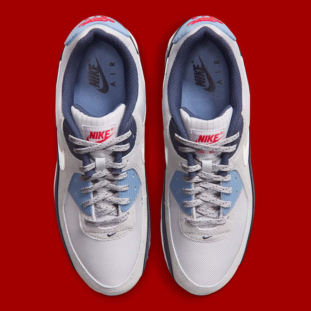 Vista superior del par de Nike Air Max 90 Geyser Grey Work Blue sobre fondo rojo, con el logo Nike rojo en la lengüeta, cordones grises con puntos, plantillas azules y gamuza gris