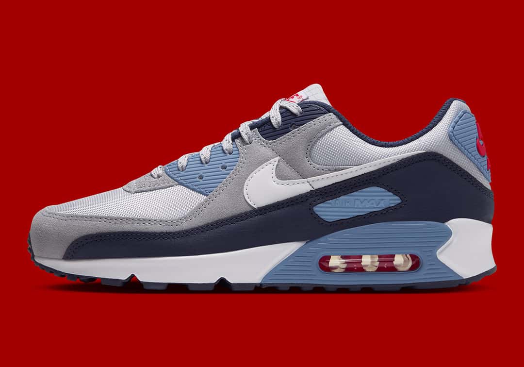 Nike Air Max 90 Geyser Grey Work Blue de perfil sobre fondo rojo, con malla gris clara, gamuza gris, puntera azul marino, Swoosh blanco, cuña Work Blue y la cámara de aire roja y blanca