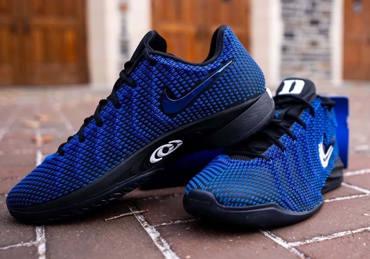 Par de Nike Caitlin 1 PE de Duke con el tejido azul sobre base negra, el Swoosh y el logo CC blancos, apoyado sobre un piso de ladrillos