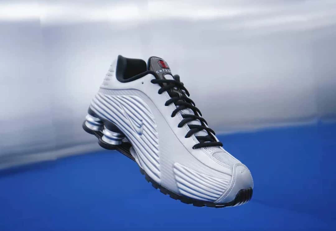 Corteiz x Nike Shox R419 plateado metalizado flotando sobre un fondo azul y plateado, con cordones negros, paneles laterales con relieves horizontales y columnas Shox metalizadas en el talón
