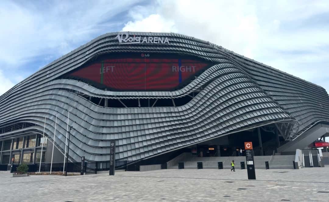 Fachada del Roig Arena de Valencia, un estadio de forma ondulada con placas grises, con una pantalla gigante en el frente y el cielo nublado de fondo