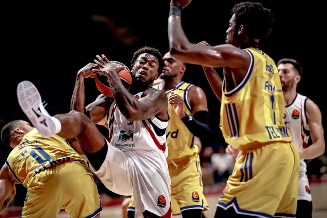 Un jugador del Olimpia Milano, con camiseta blanca, se lleva la pelota entre tres jugadores del Maccabi Tel Aviv de camiseta amarilla, uno de ellos caído en el piso