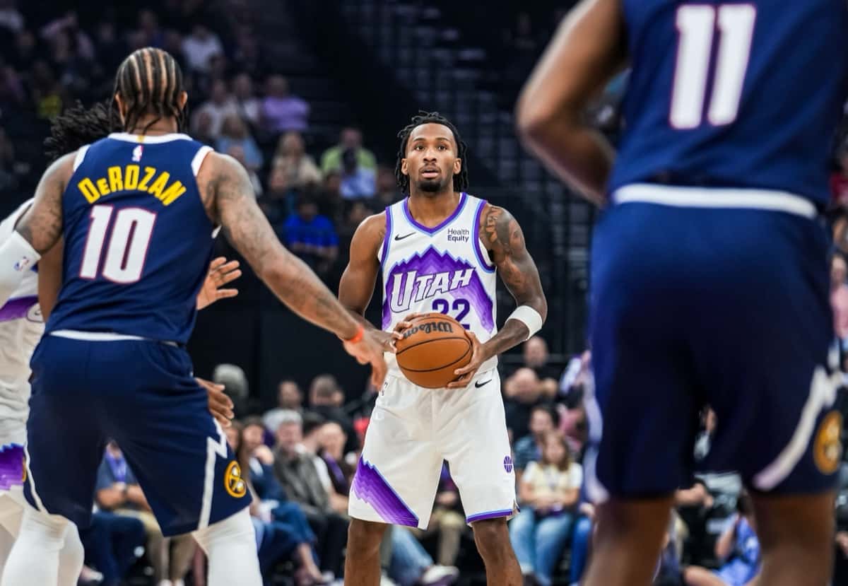 Darryn Peterson, con la camiseta blanca número 22 del Utah Jazz, sostiene la pelota frente a la marca de dos jugadores de los Denver Nuggets vestidos de azul
