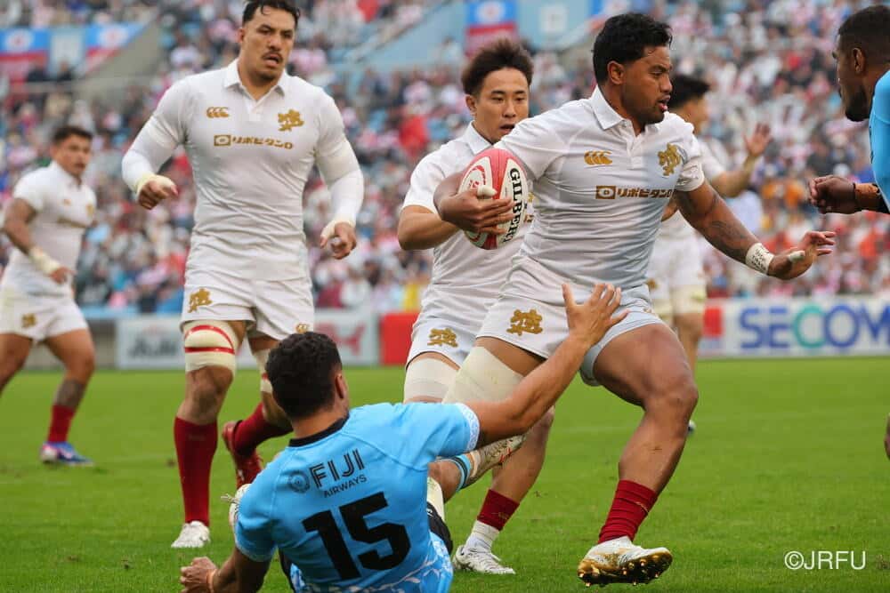 Un forward de Japón, con la camiseta blanca, avanza con la pelota ante el fullback de Fiyi, de celeste, en el test del estadio Chichibunomiya