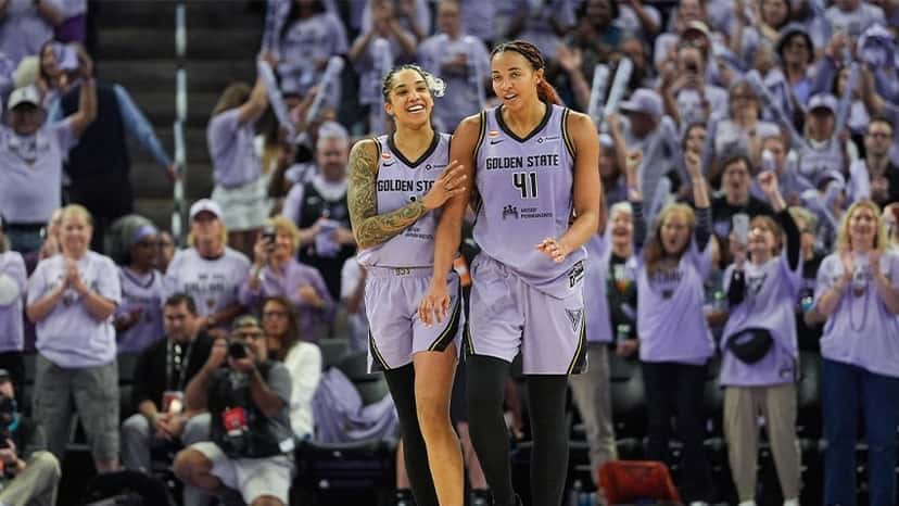 Gabby Williams, con la camiseta lila de Golden State Valkyries, sonríe abrazada a Kiah Stokes, con la 41, mientras la tribuna del Chase Center festeja de pie