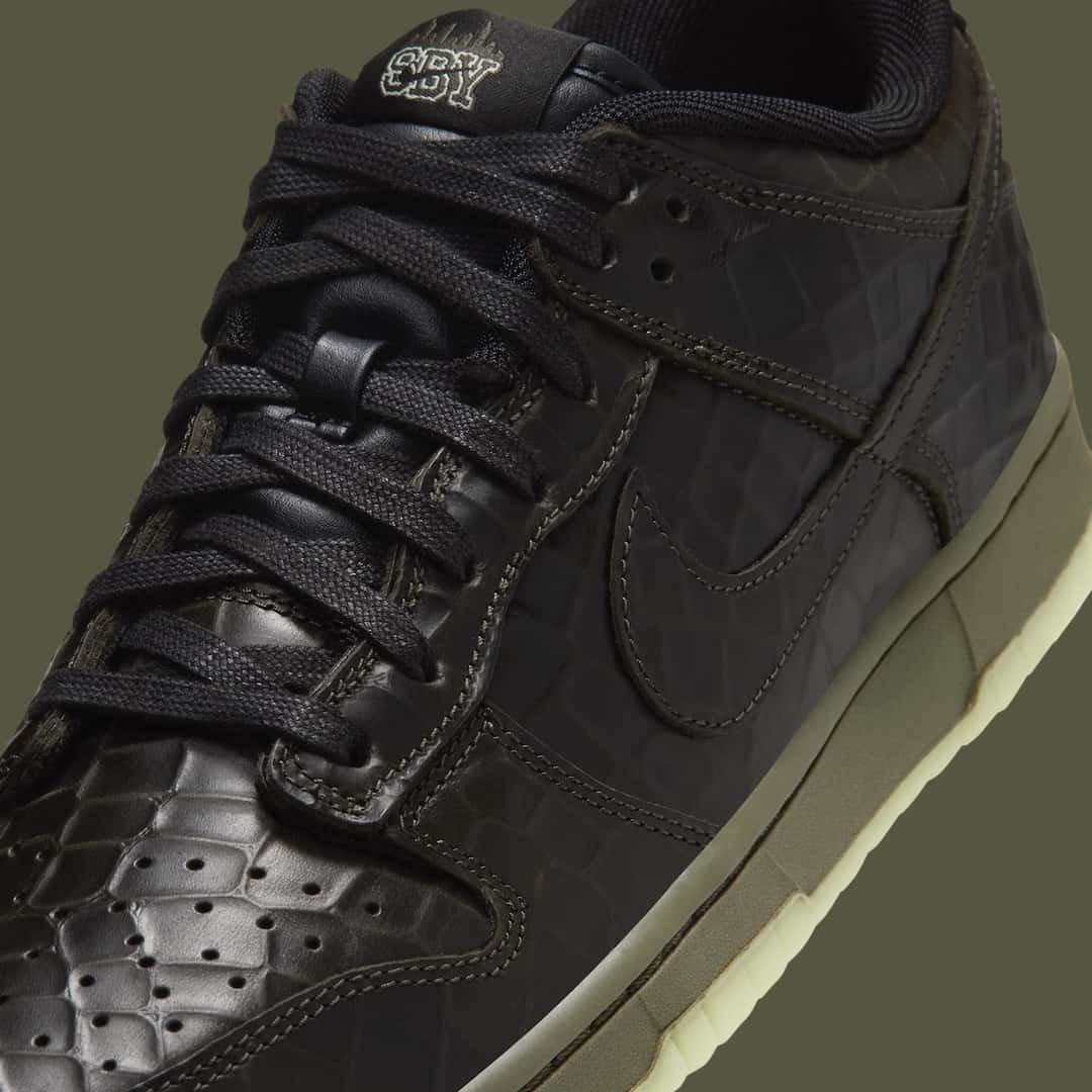 Primer plano del Nike Dunk Low Shibuya Halloween con cordones negros, cuero negro texturado tipo cocodrilo y la sigla SBY bordada en la etiqueta de la lengüeta