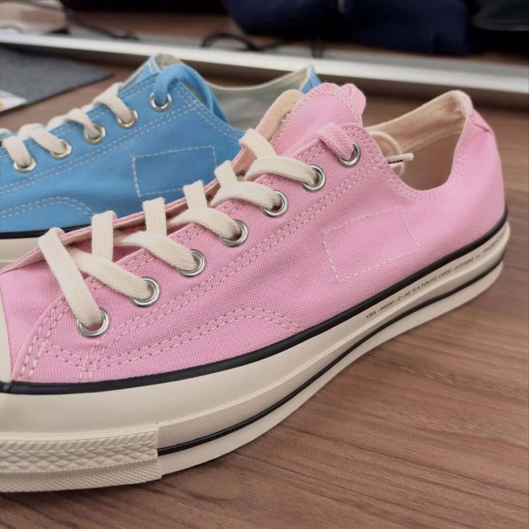 Converse Chuck Taylor baja de fragment design en rosa, de perfil sobre una mesa de madera, con un par celeste detrás y una costura rectangular en la capellada