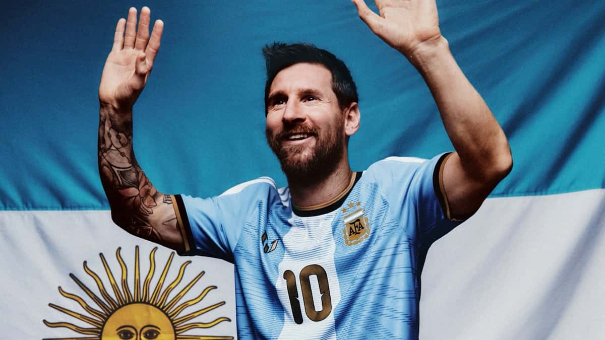 Lionel Messi saluda con las dos manos en alto vestido con la camiseta especial de su despedida, celeste y blanca con el 10 dorado, delante de una bandera argentina (foto oficial de adidas)
