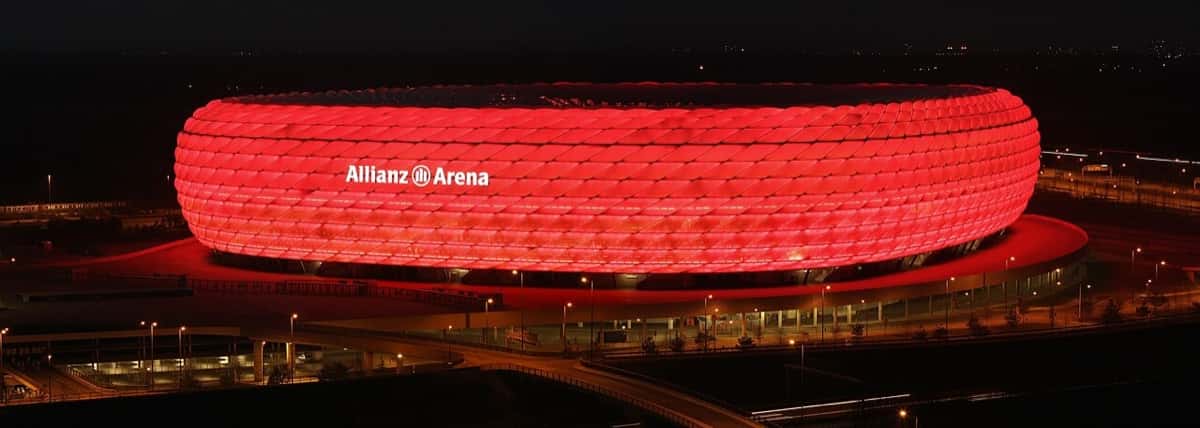 El Allianz Arena de Múnich iluminado de rojo por la noche, con el estacionamiento y la explanada alrededor; foto de archivo de enero de 2008 (foto: Richard Bartz, Wikimedia Commons, licencia CC BY-SA 2.5)
