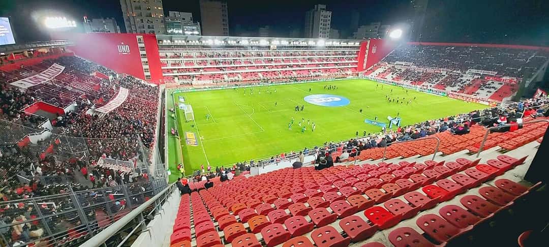 Vista nocturna del estadio UNO Jorge Luis Hirschi, la cancha de Estudiantes en La Plata, con las tribunas rojas y blancas y el campo iluminado (foto de archivo: Germanramos2 / Wikimedia Commons, CC BY-SA 4.0)