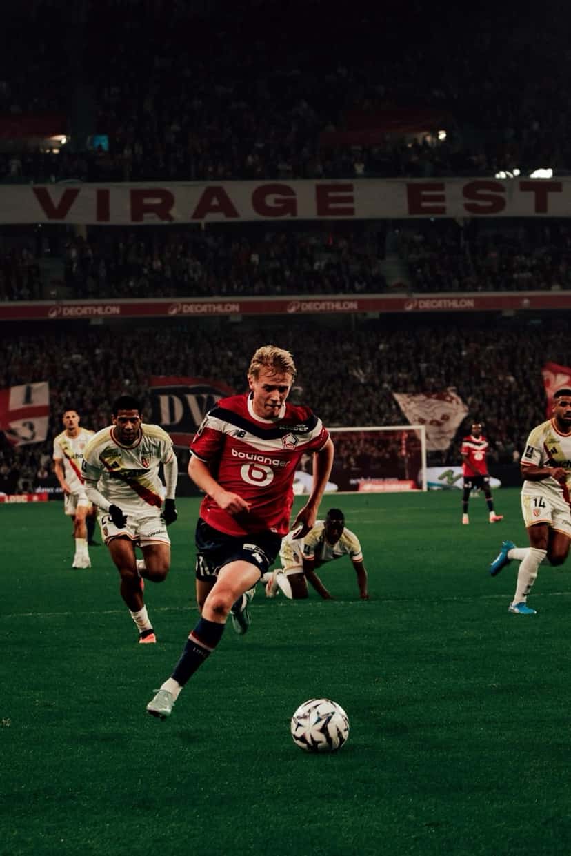 Un jugador del Lille, con la camiseta roja, conduce la pelota perseguido por jugadores de Lens de camiseta blanca, con la tribuna local de fondo, el viernes 30 de octubre (foto: LOSC)