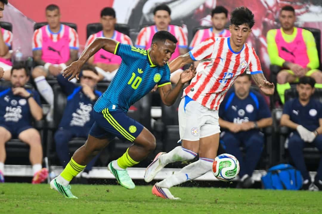 Un jugador de Colombia con la camiseta 10 y un jugador de Paraguay disputan la pelota junto al banco paraguayo en el Sports Illustrated Stadium