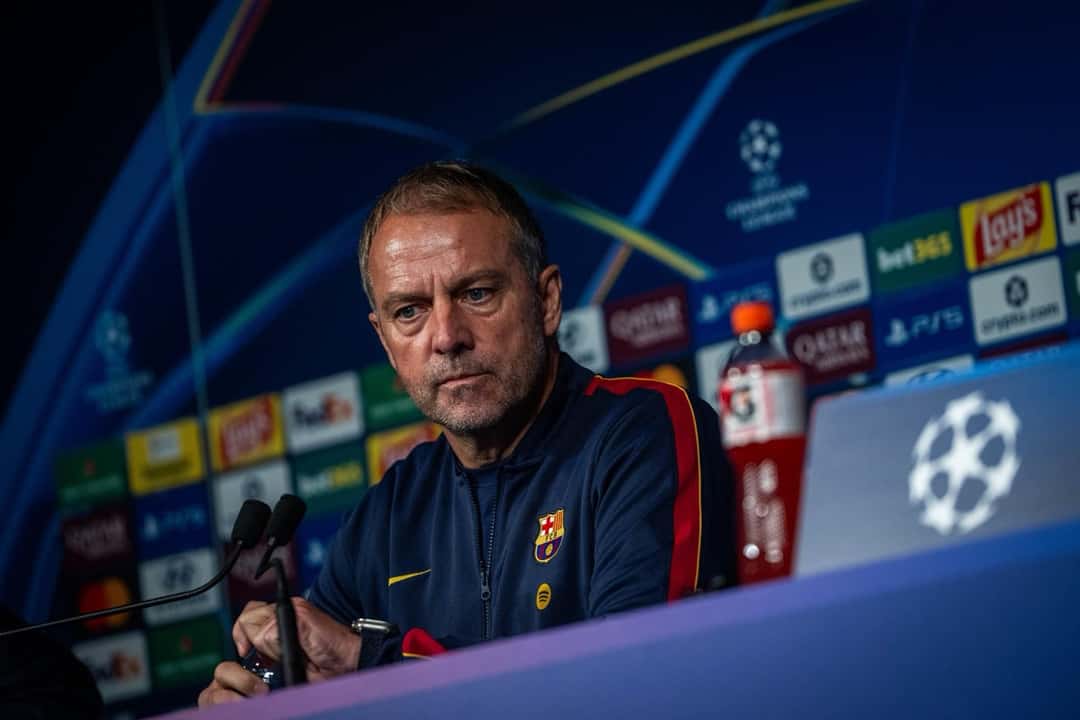 Hansi Flick, entrenador del Barcelona, con campera azul oscuro del club, en la conferencia de prensa previa al partido de Champions con el PSG, frente a un panel con el logo del torneo, el lunes 19 de octubre en París (foto: FC Barcelona)
