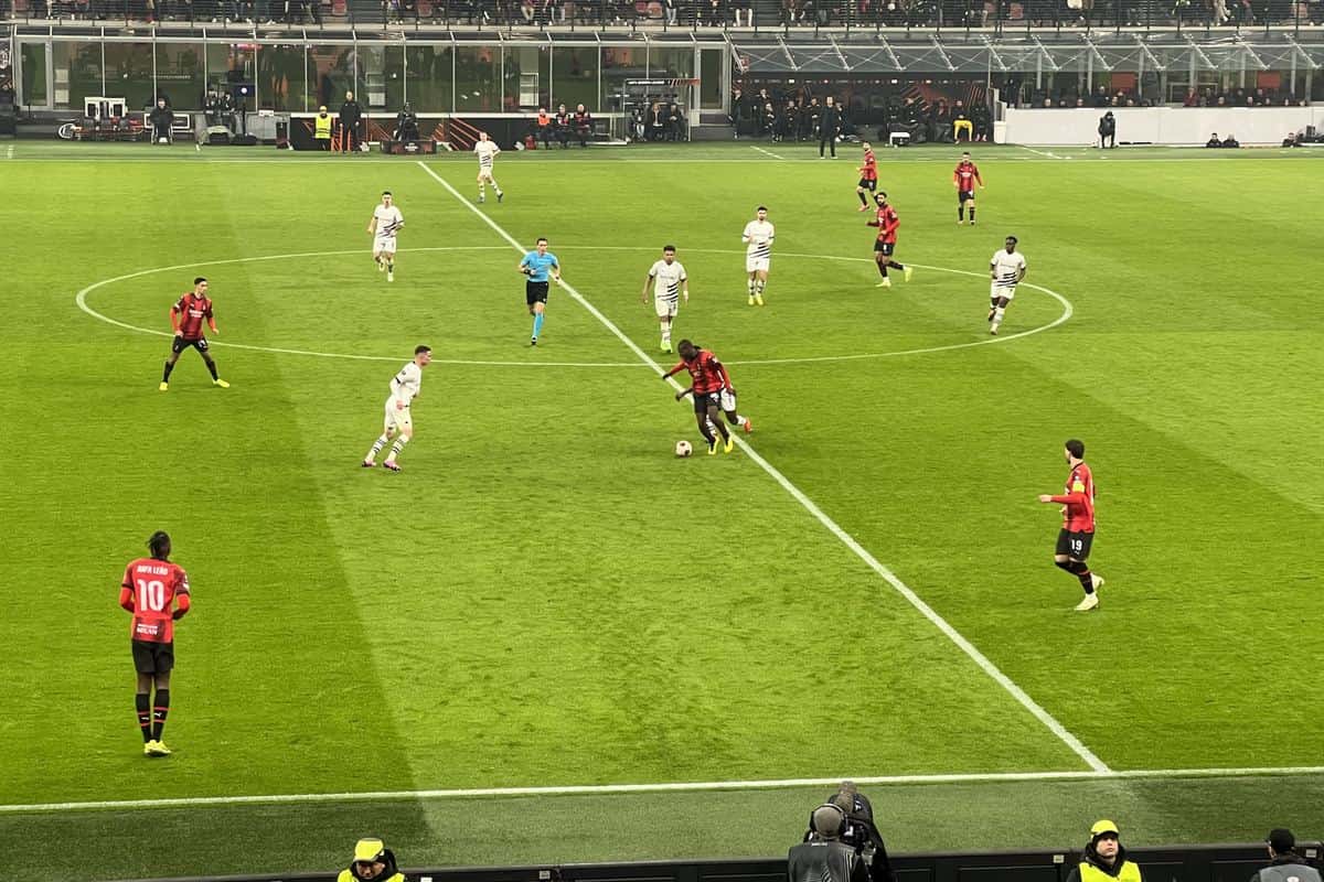 Un partido del Milan en el estadio San Siro, con jugadores de camiseta roja y negra y rivales de blanco en el círculo central y la tribuna detrás; foto de archivo de febrero de 2024 (foto: LHC88, Wikimedia Commons, licencia CC BY 4.0)