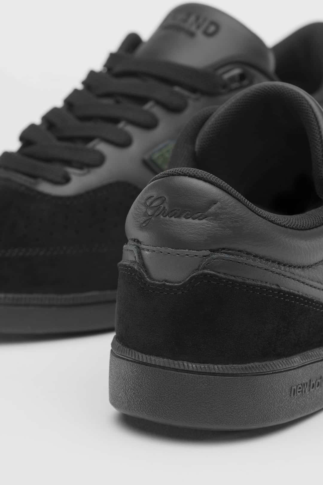 Primer plano del talón de la New Balance Numeric 508 de Grand Collection en negro, con la palabra Grand grabada en letra cursiva sobre el cuero y el logo de New Balance en relieve en la suela