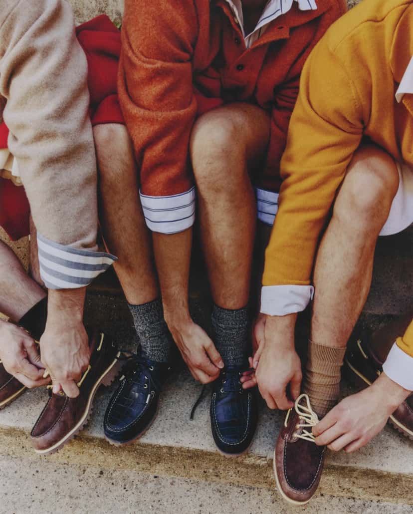 Foto de campaña con tres personas sentadas en un escalón atándose los Timberland x Jacquemus Authentic Boat Shoe: dos con pares dark brown de cordones amarillos y una con el par navy en relieve de cocodrilo, con medias grises y beige y suéteres naranja y amarillo