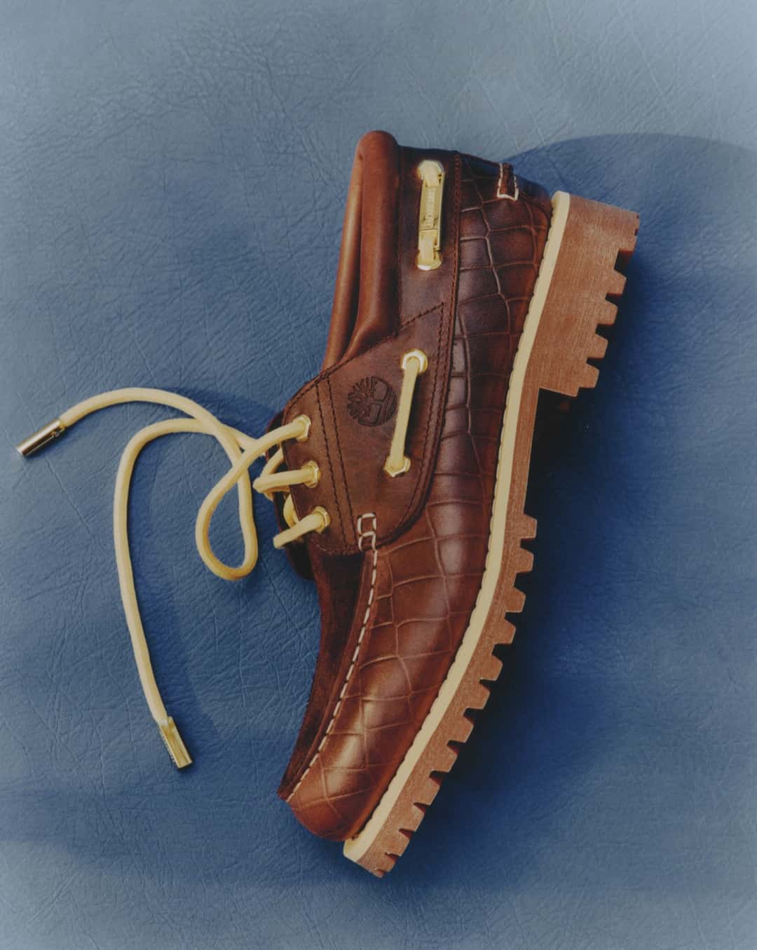 Zapato Dark Brown del Timberland x Jacquemus Authentic Boat Shoe de perfil sobre un fondo de cuero azul, con el cuero marrón en relieve de cocodrilo en la parte baja, el árbol de Timberland grabado en el lateral, pasacordones y herretes dorados, cordones amarillo claro y la suela de goma con tacos en tono natural
