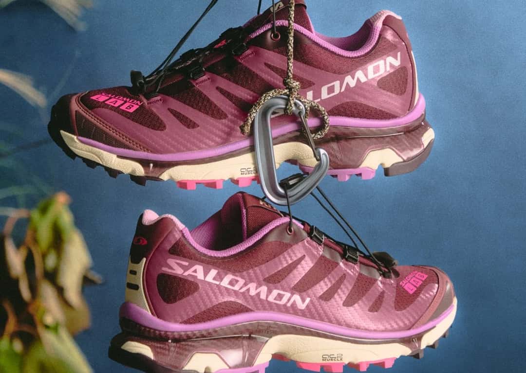 Par de Kith x Salomon XT-4 OG en bordó con detalles rosa colgando de un mosquetón sobre fondo azul
