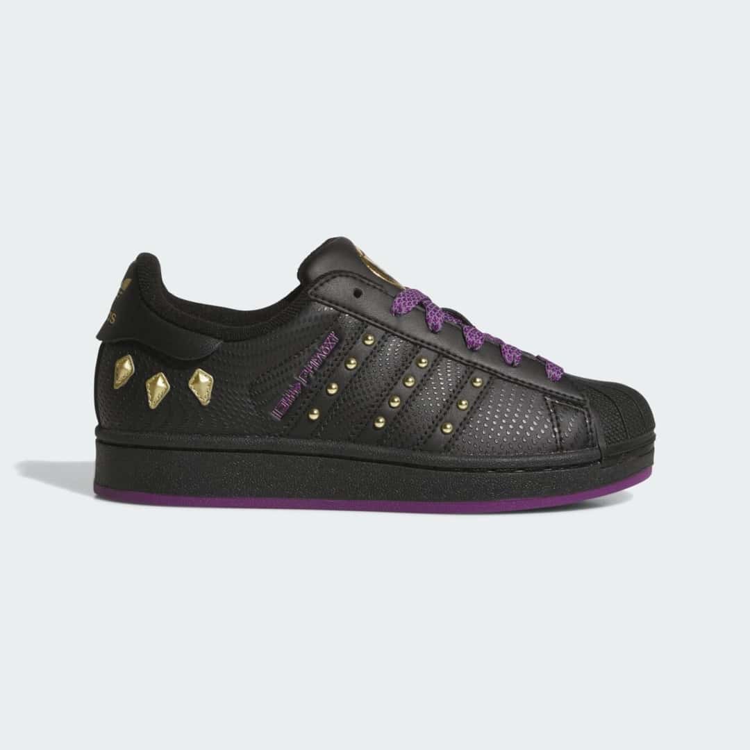 adidas Superstar Shuri Black Panther de perfil en cuero negro texturado, con tachas doradas en las tres tiras, piezas doradas en el talón, cordones violetas y suela violeta