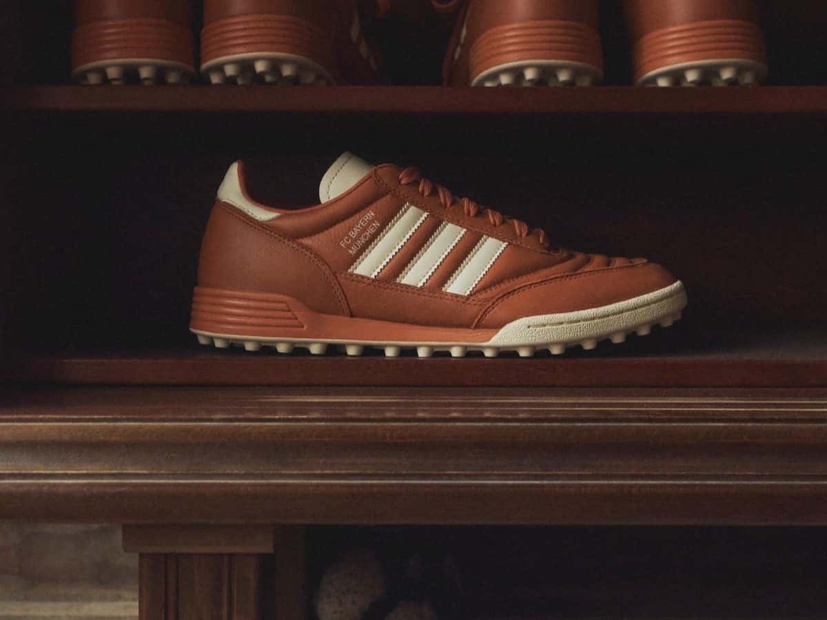 Zapatilla adidas Mundial Team en cuero color coñac con las tres tiras en crema y suela de tapones cortos, sobre un estante de madera
