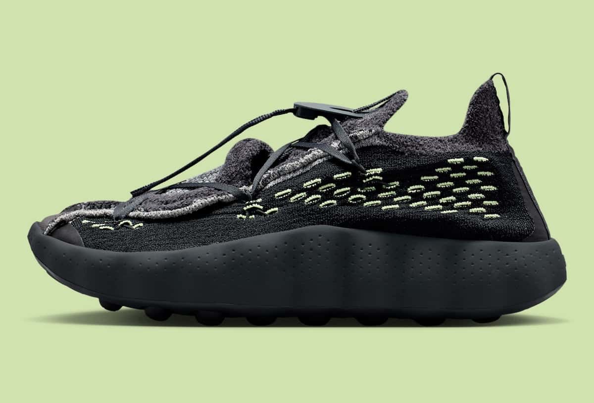 Nike Mind 002 Flyknit Black/Dark Smoke Grey de perfil, con tejido negro y gris, costuras verde lima y suela negra, sobre fondo verde claro